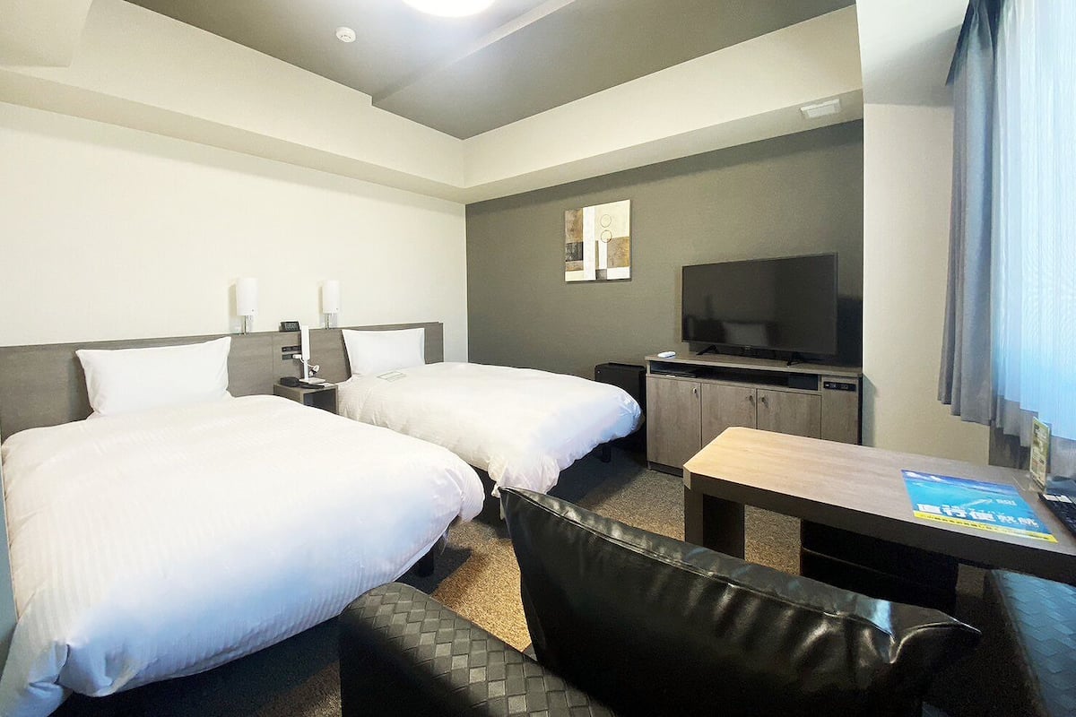 Ein Hotelzimmer mit zwei Betten, einem Schreibtisch, einem Fernseher und einem Fenster mit Vorhängen.