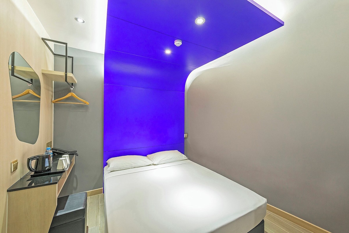 A modern hotel room with a bed, a mirror, a small table with a kettle, and a blue accent wall.