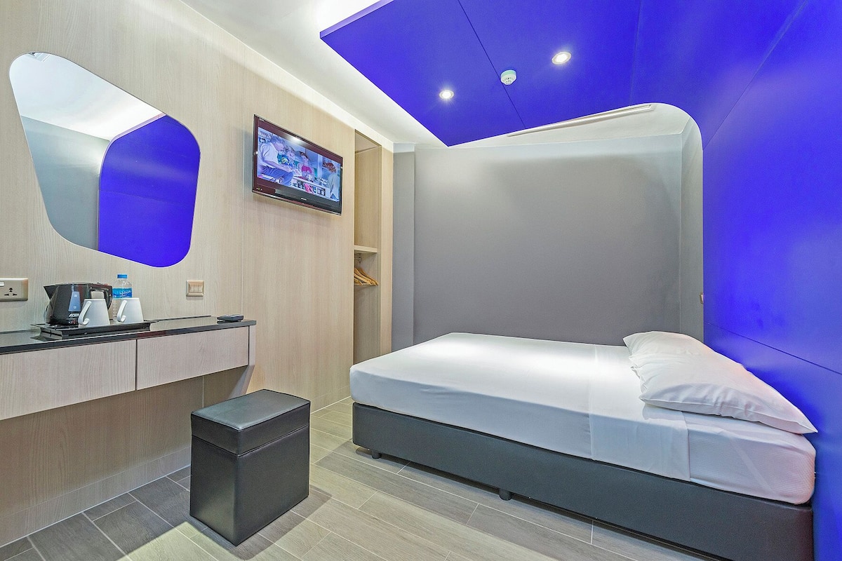 A modern hotel room with a bed, a desk with a coffee maker, a TV, and a small trash bin.