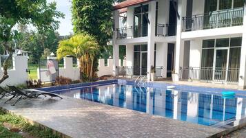 Piscina com deck revestido, rodeada por vegetação e um edifício de dois andares com sacadas.
