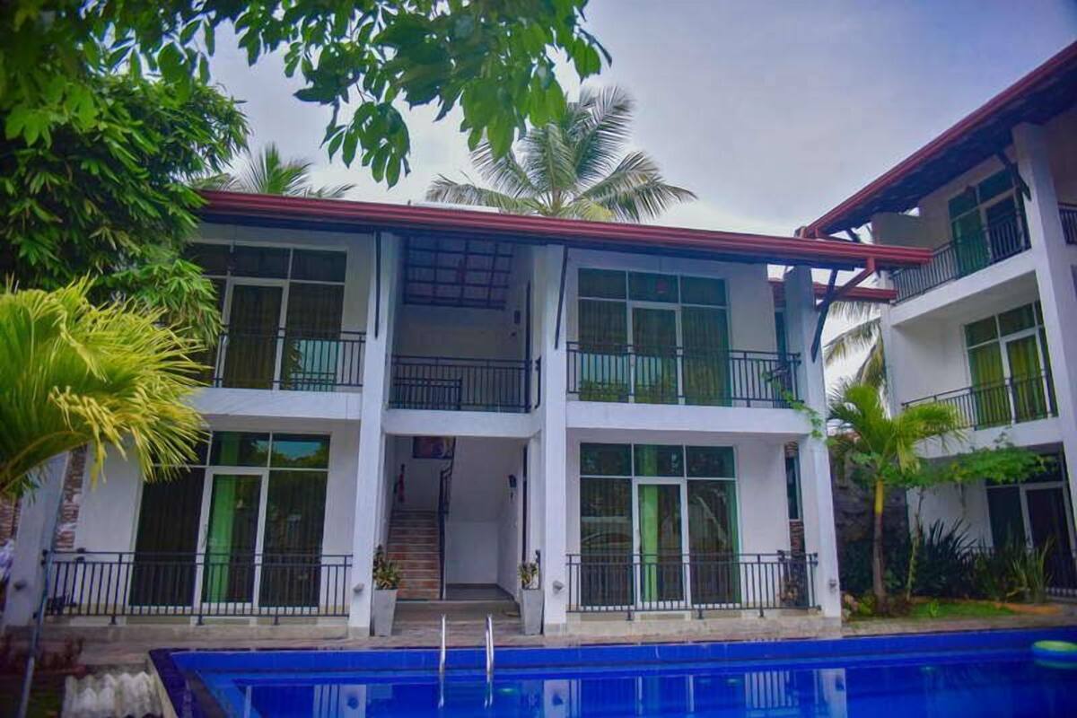 Un edificio de dos plantas con piscina delantera, rodeado de vegetación.