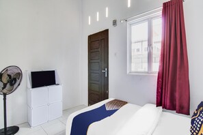 Une pièce comprenant un lit, un ventilateur sur pied, un meuble de rangement blanc, une porte et une fenêtre avec des rideaux.