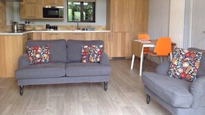 A modern kitchen with wooden cabinets and a microwave, a grey sofa with colorful cushions, and a dining area with a table and chairs.