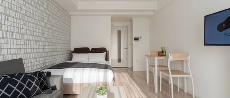 A modern bedroom with a bed, a grey sofa, a small table with a plant, and a dining area with a chair and table.