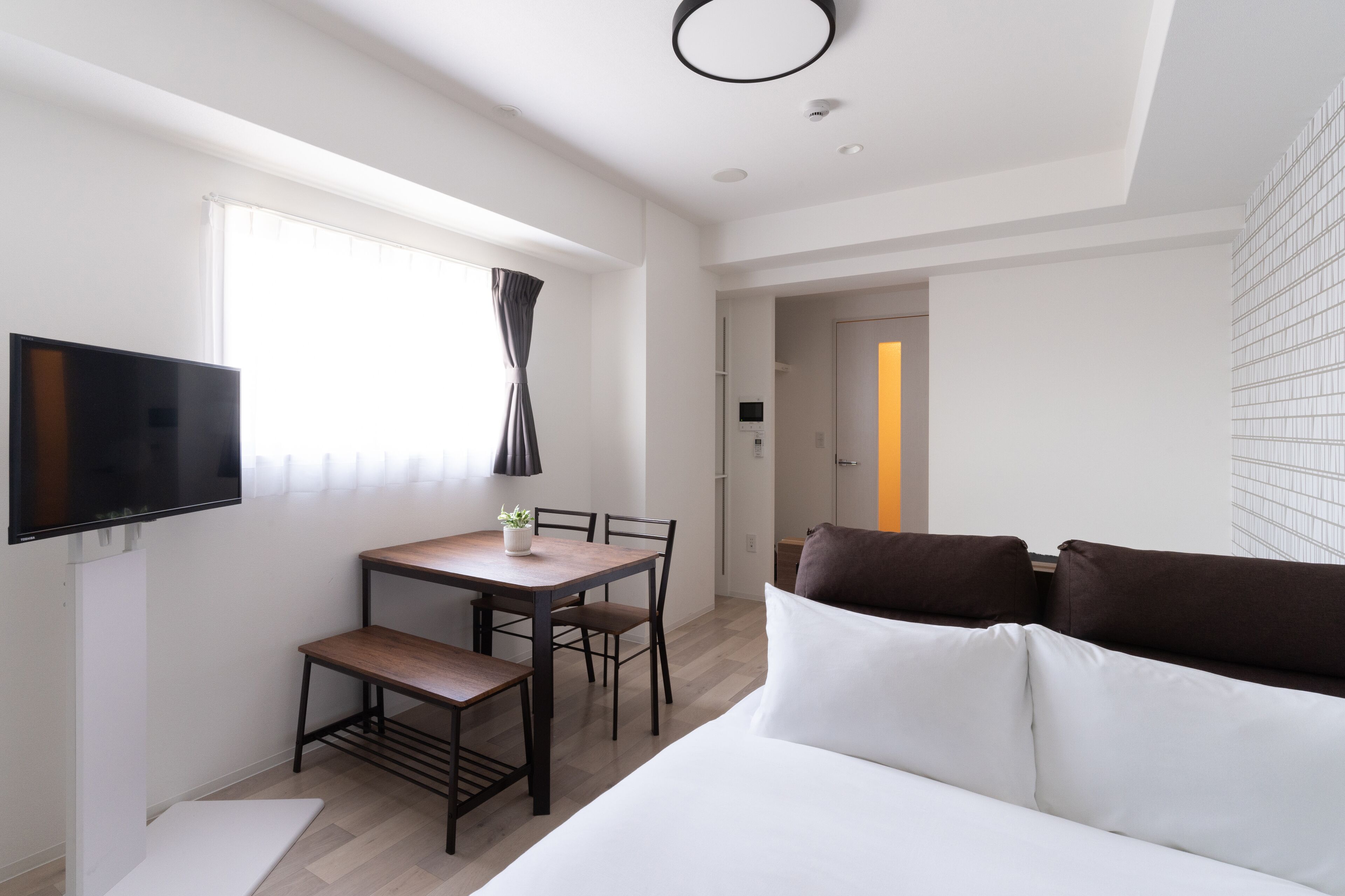 A modern hotel room with a bed, a TV, a dining table with chairs, and a small plant.