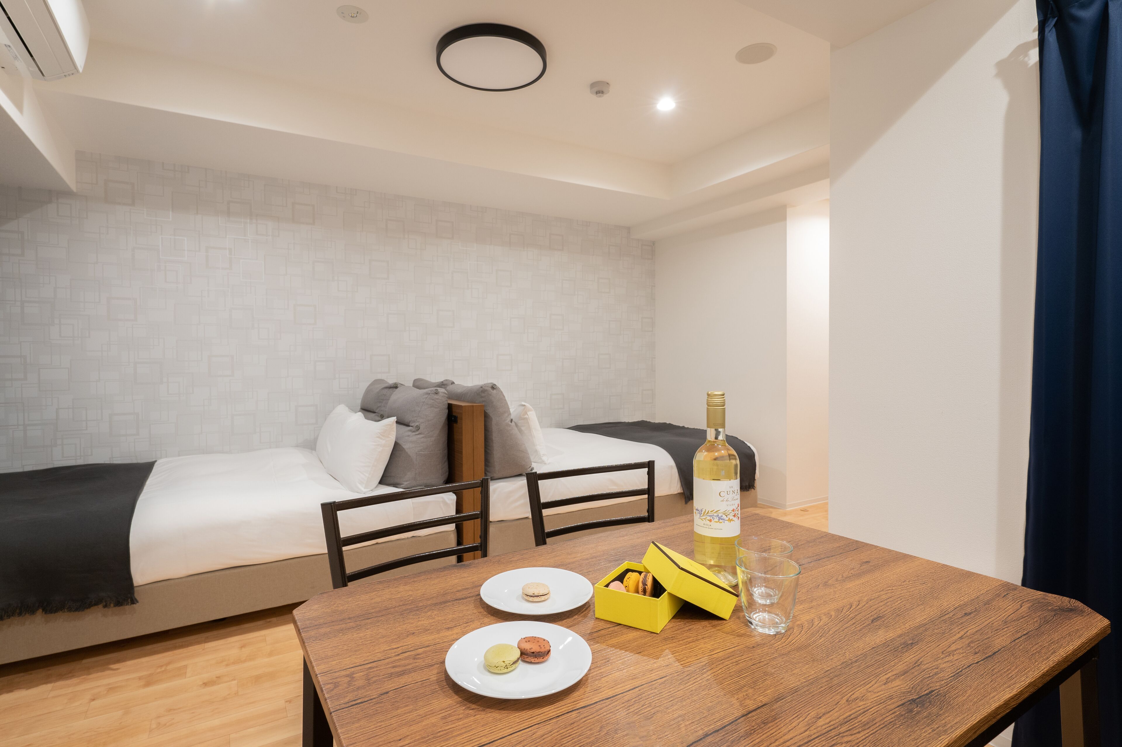 A hotel room with a bed, a wooden table with a bottle and glasses, and a box of macarons.