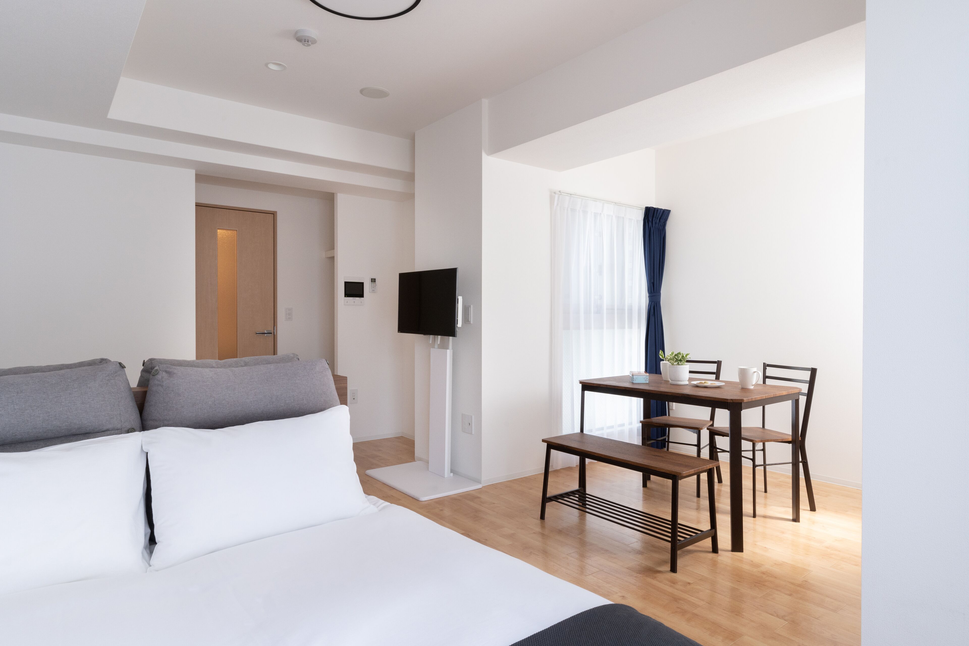 A modern hotel room with a bed, a TV, a dining area with a table and chairs, and a wooden floor.