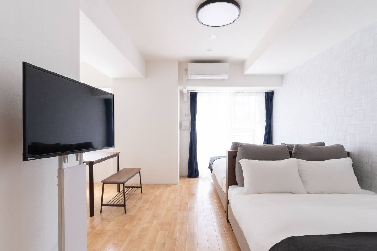 Een moderne hotelkamer met een flatscreen-tv, een houten bankje, een bed met wit en grijs beddengoed, en een raam met gordijnen.