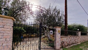 A gated entrance with a stone wall and metal gate, leading to a park area with trees and a bench. - Villa Kavalino (Κολύμπια)