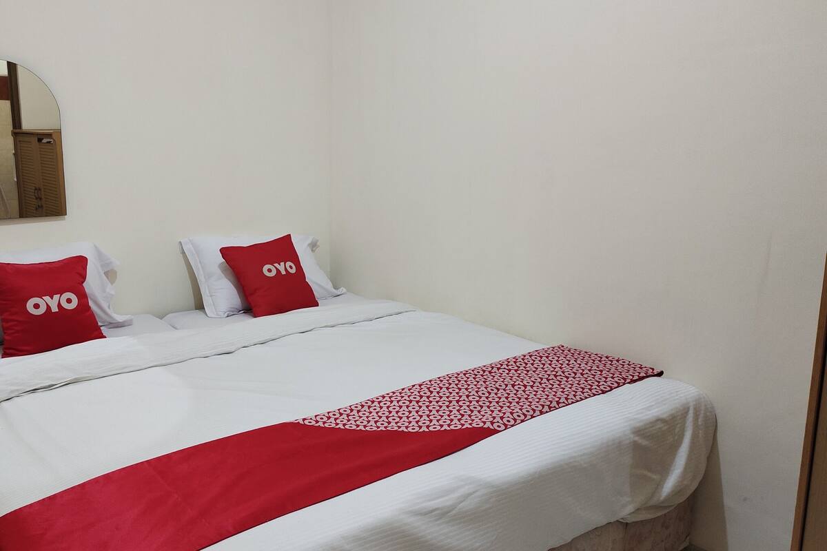 Una habitación de hotel con una cama, dos almohadas con la marca 'OYO', una colcha roja y blanca, y un espejo en la pared.