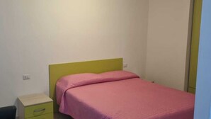 A small, single-bed room with a green headboard and a pink bedspread. There is a small bedside table with a drawer and a lamp on the wall. - Piccolo Apartment for 2 people (Budoni)