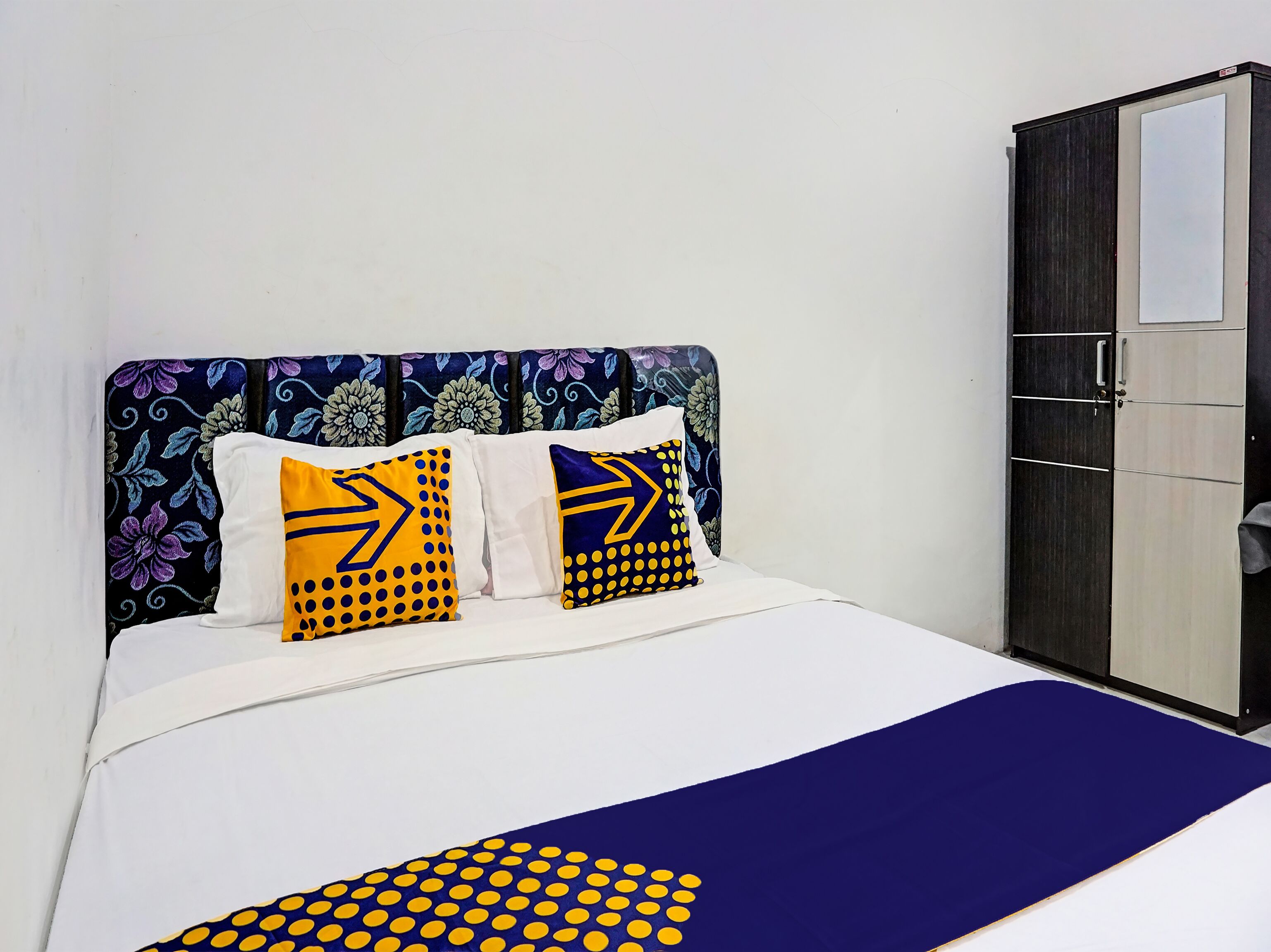 A bed with a white and blue bedspread, two pillows with geometric patterns, and a dark wardrobe against a plain wall.