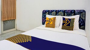 A bed with a white and blue bedspread, two pillows with yellow and blue patterns, a floral headboard, and a window with brown curtains. - OYO Life 91718 Kost Damai Syariah (Sidoarjo)