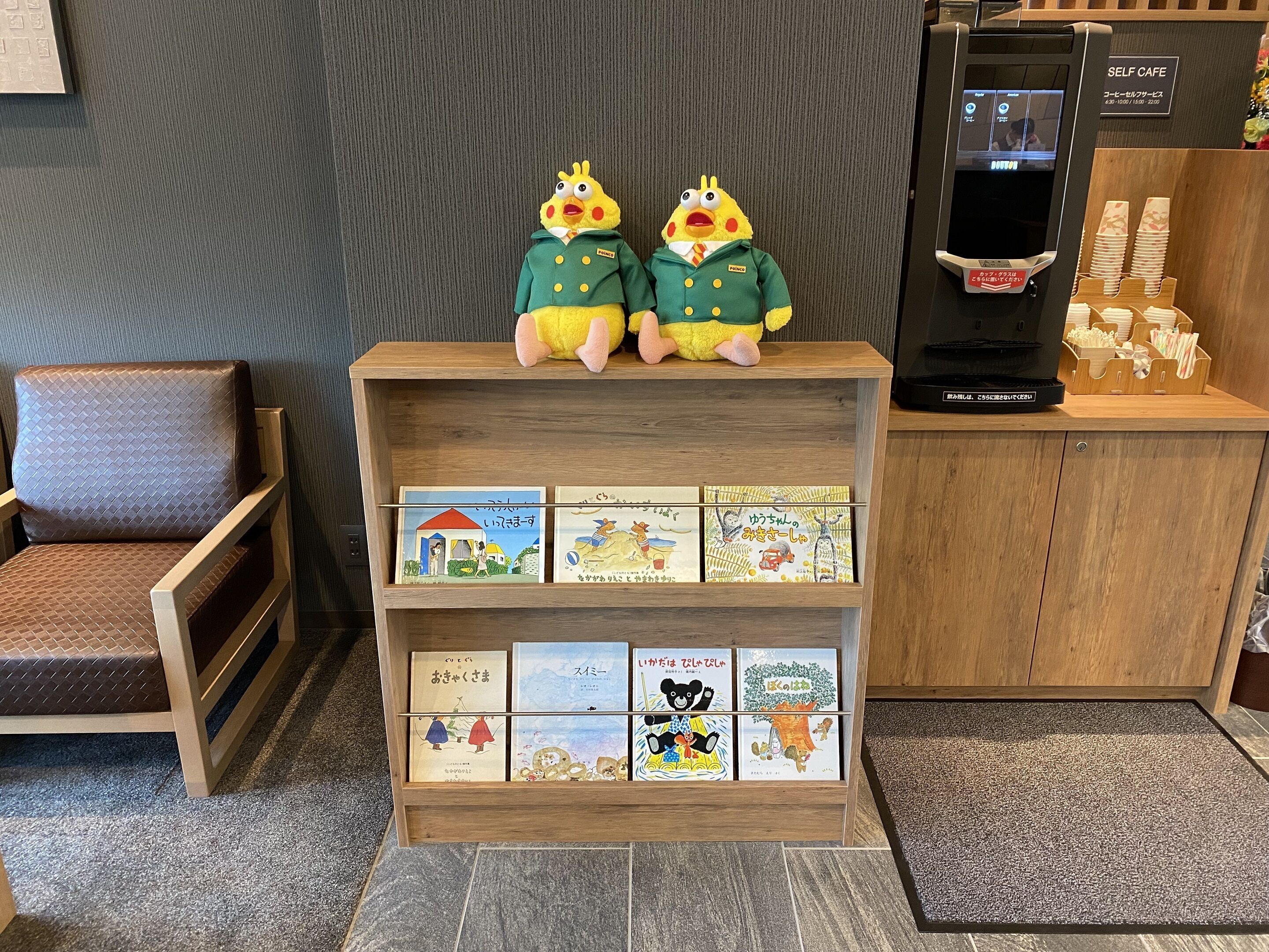 Una máquina expendedora de café, un peluche y un estante de madera con libros infantiles.