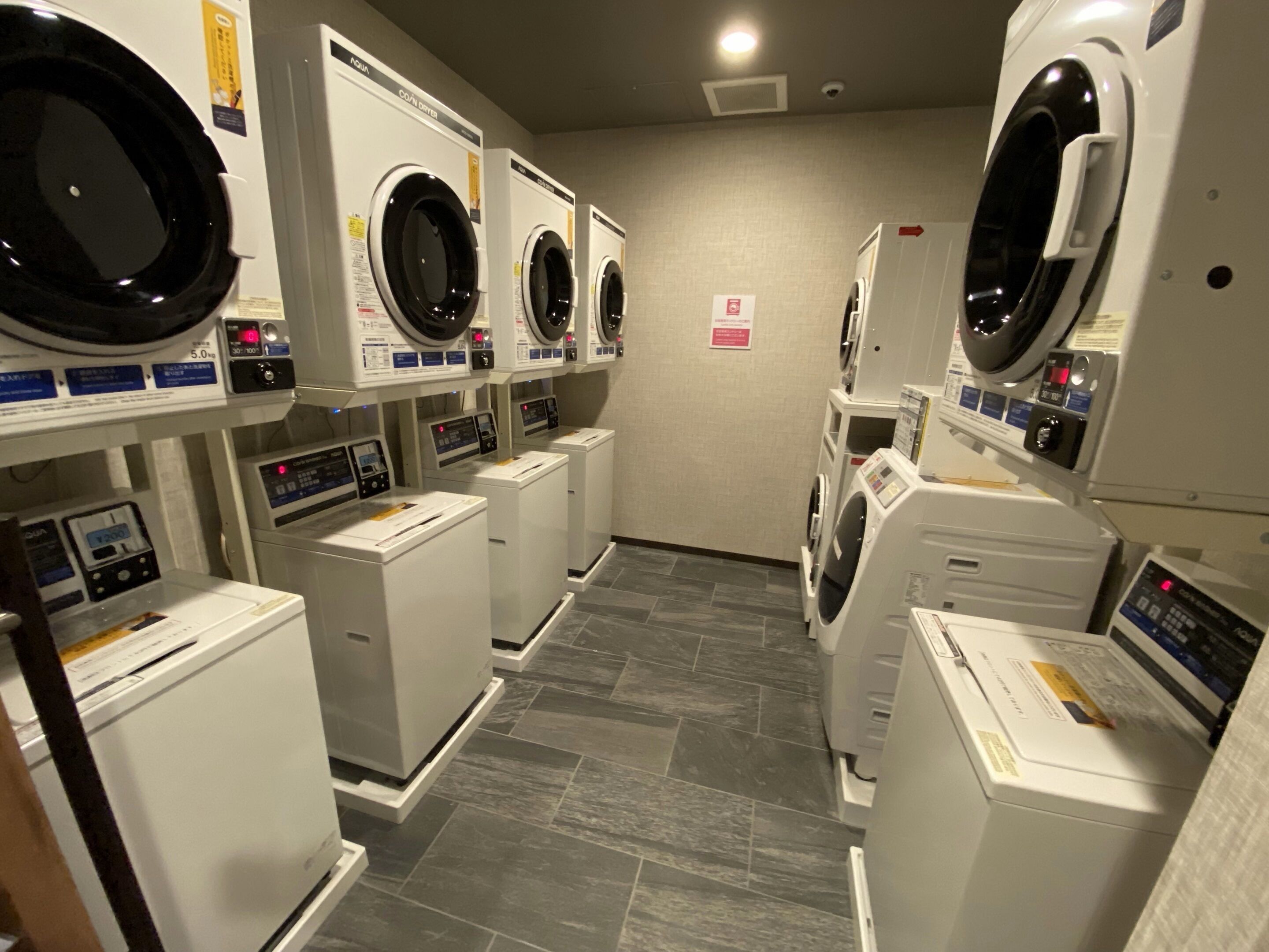 A laundry facility with multiple washing machines and dryers, arranged in a row with a tiled floor.