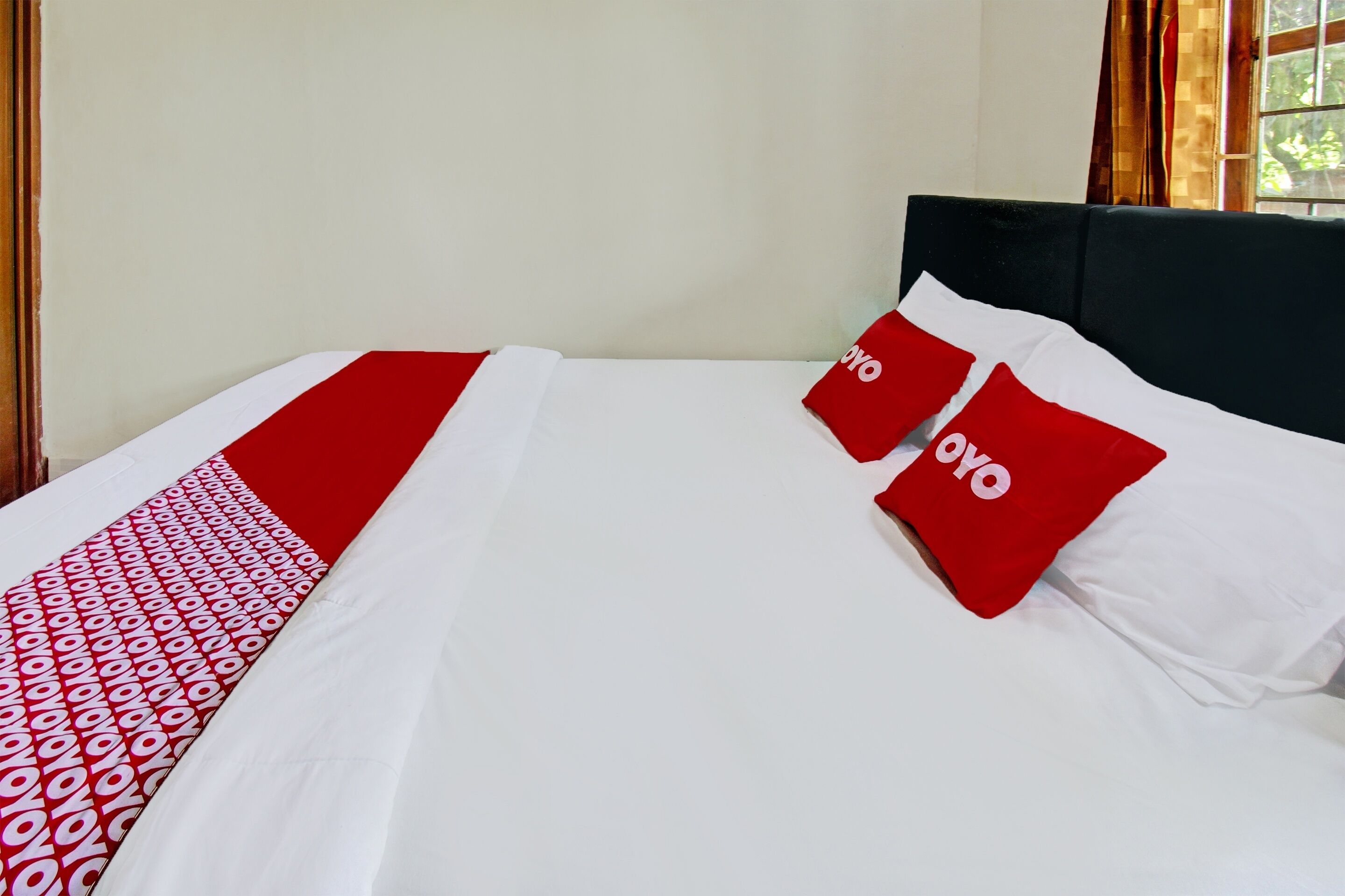 A neatly made bed with a white and red bedspread, two red pillows with the word "oyo" on them, and a window with curtains in the background.