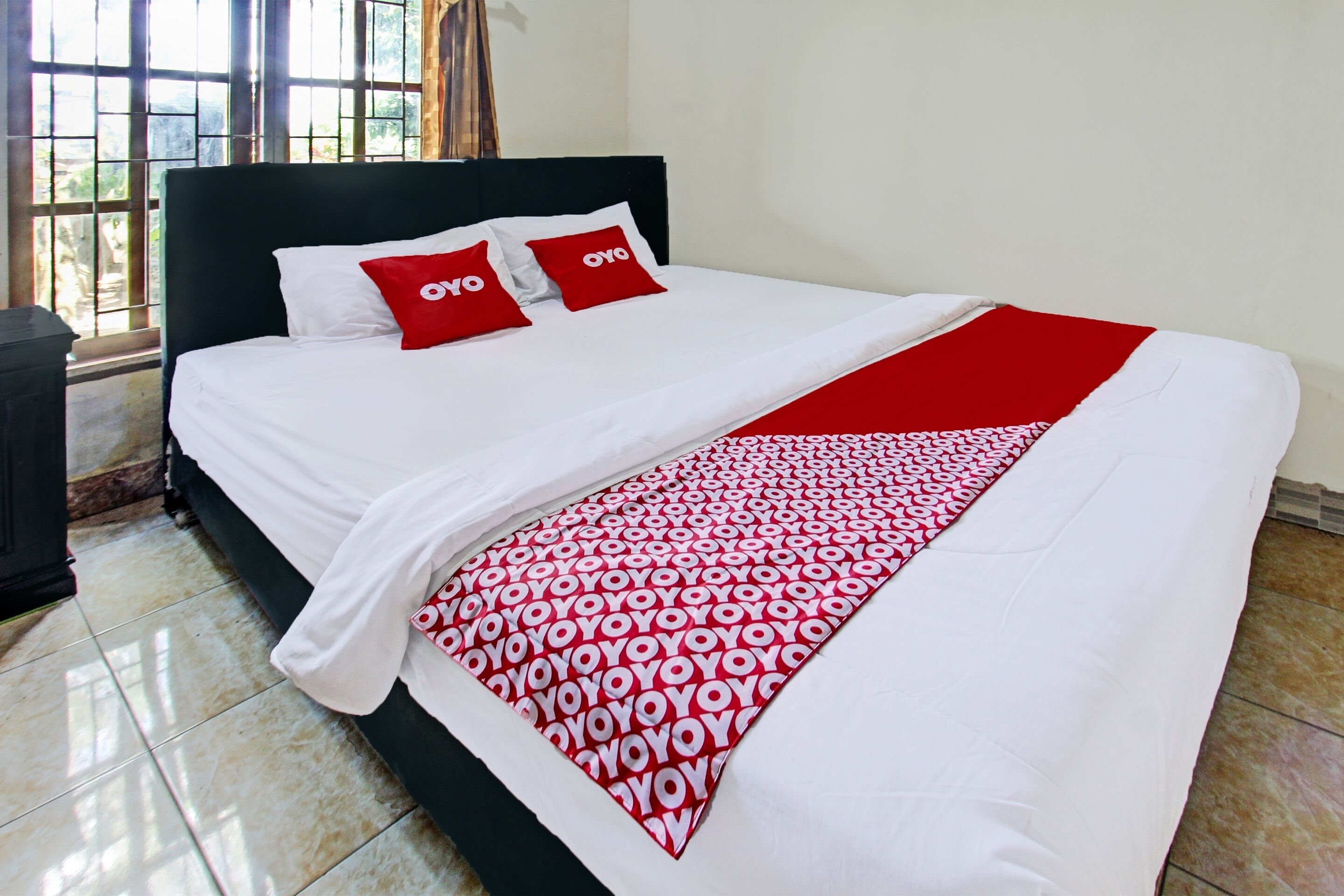 Una cama con ropa de cama blanca y detalles rojos, un plaid rojo estampado y una ventana con vista a follaje.
