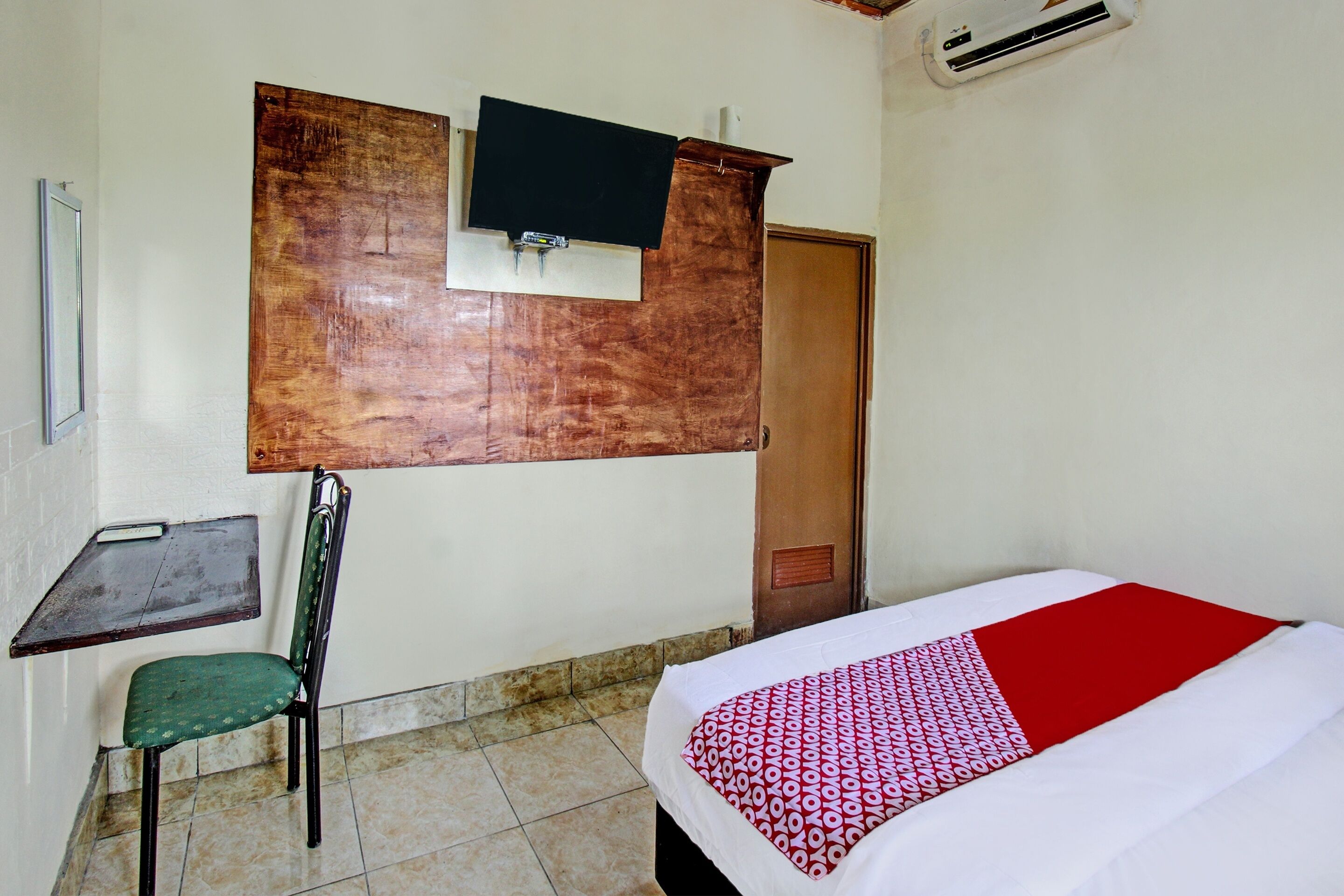 A room with a bed, a chair, a desk, a television mounted on the wall, and an air conditioning unit.