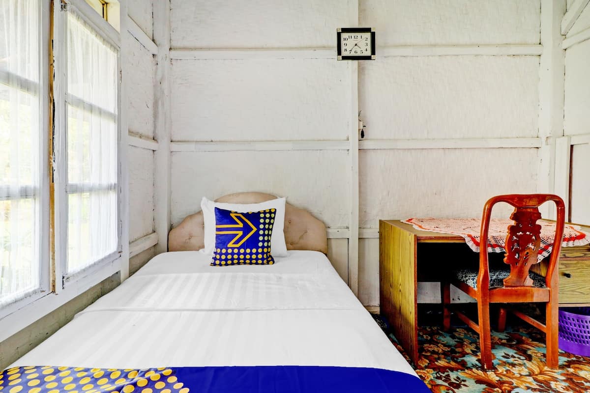 A small, single-bed room with a wooden headboard, a bedside table with a red chair, and a window with curtains.