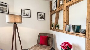 Una habitación con una silla, una lámpara de pie, una mesita con un florero y una estantería de madera con libros y adornos.