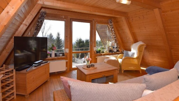 Een gezellige zolderkamer met houten wanden en een schuine kap. Er is een televisie, een houten kast, een bank en een stoel.