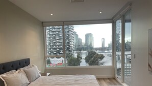 A modern bedroom with a large bed, a view of a cityscape through floor-to-ceiling windows, and a sliding glass door leading to an outdoor balcony. - Luxury Penthouse Above the Shopping Town (Docklands)