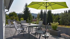 An outdoor patio with a table and chairs, a green umbrella, and a view of trees and hills.