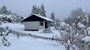 A small, single-story house with a dark roof and white walls, surrounded by snow-covered trees and a wooden fence.