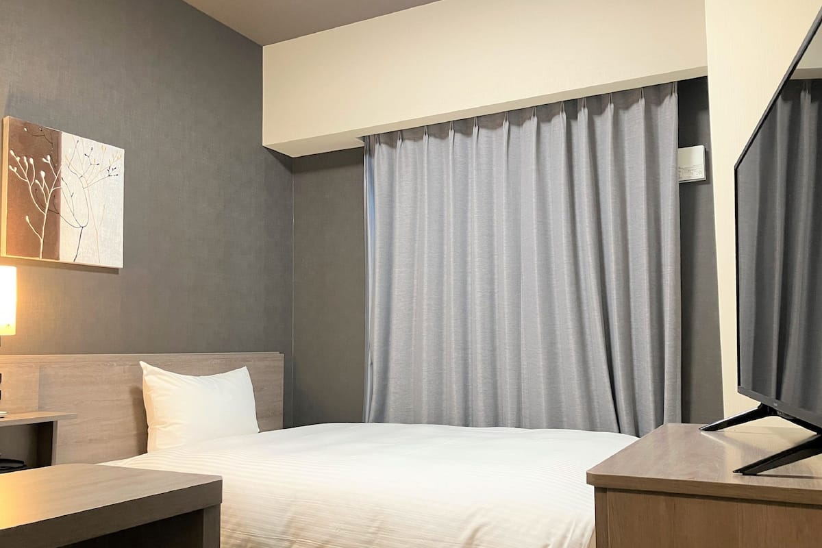 A hotel room with a bed, a desk, a chair, a TV, and a window with curtains.