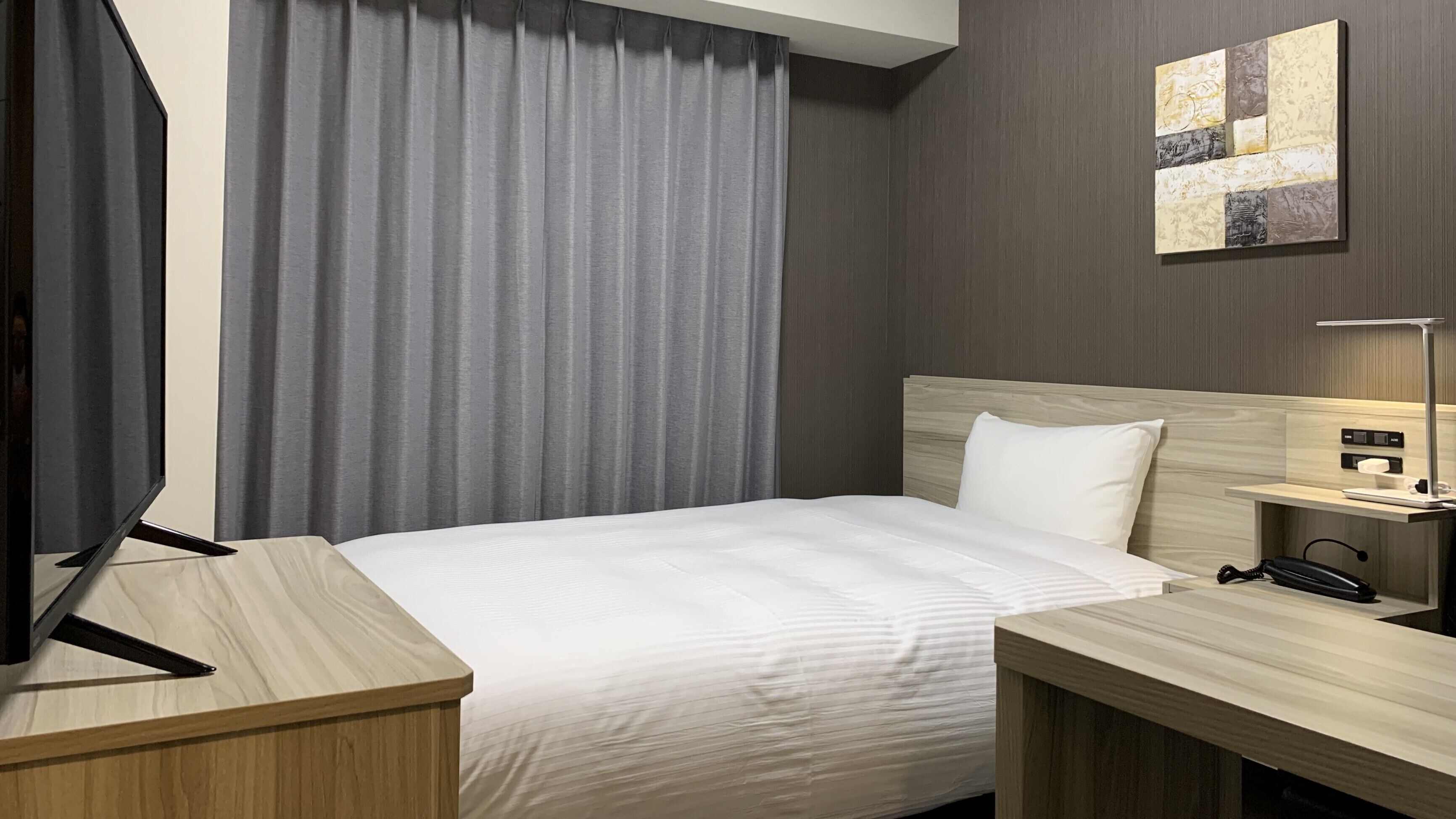 A hotel room with a bed, a desk, a TV, a lamp, and a painting on the wall.