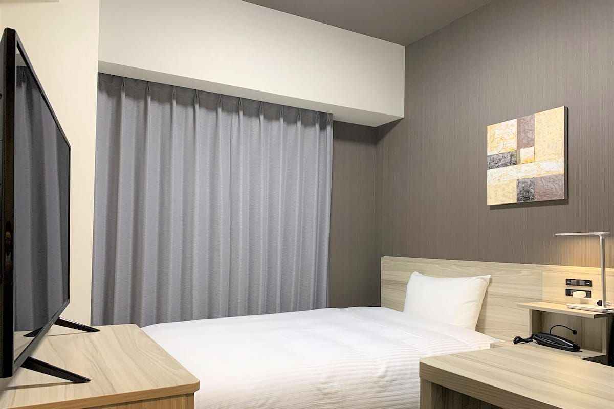 A hotel room with a bed, a desk with a telephone, a TV, and a painting on the wall.