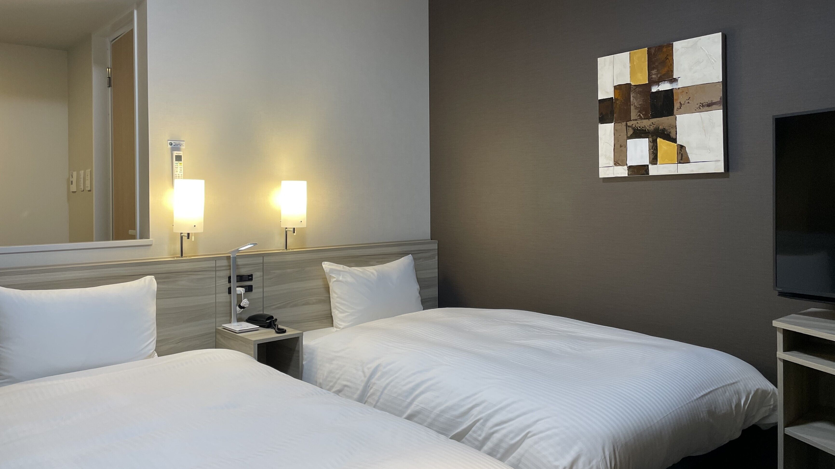 A hotel room with two beds, a nightstand, a lamp, a telephone, a flat-screen TV, and a wall-mounted artwork.
