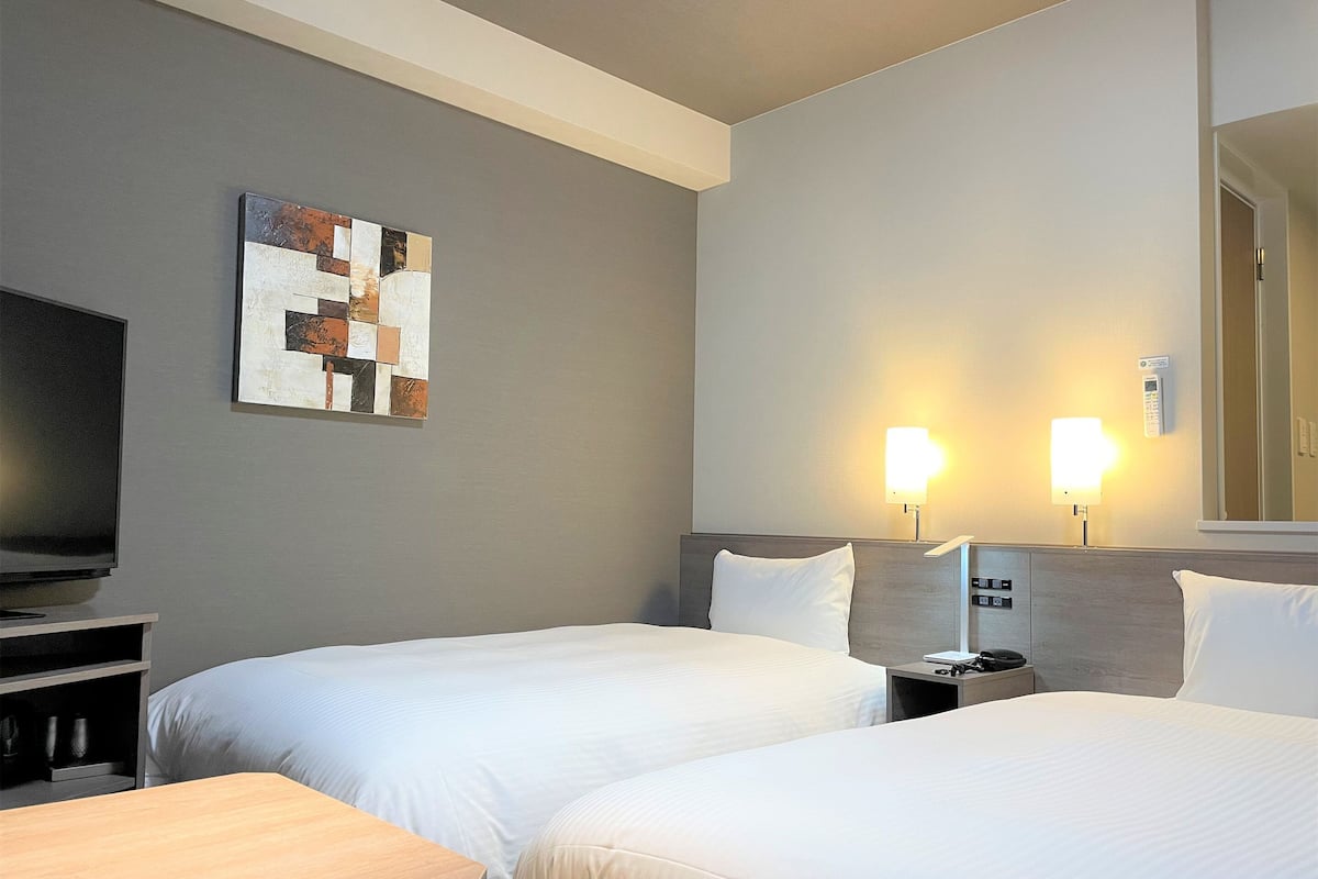 A hotel room with two beds, a television, bedside lamps, a painting on the wall, and a wooden bench.