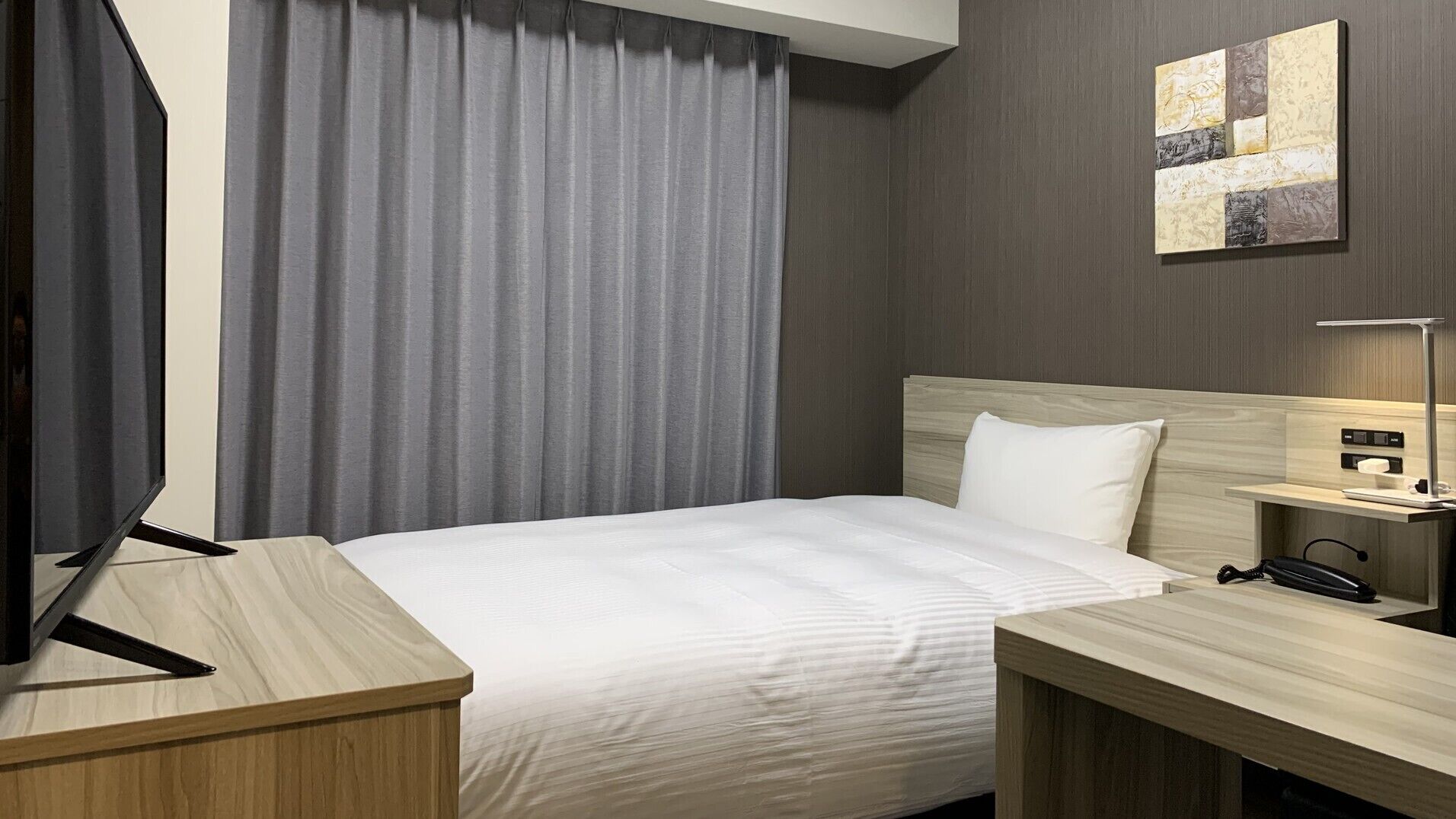 A hotel room with a bed, a desk, a TV, a bedside table with a lamp, and a wall art piece.