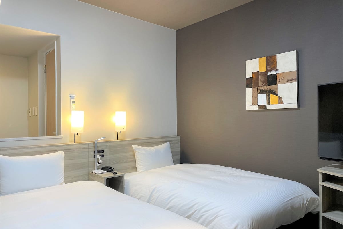 A hotel room with two beds, a nightstand with a lamp, a wall-mounted TV, and a painting.