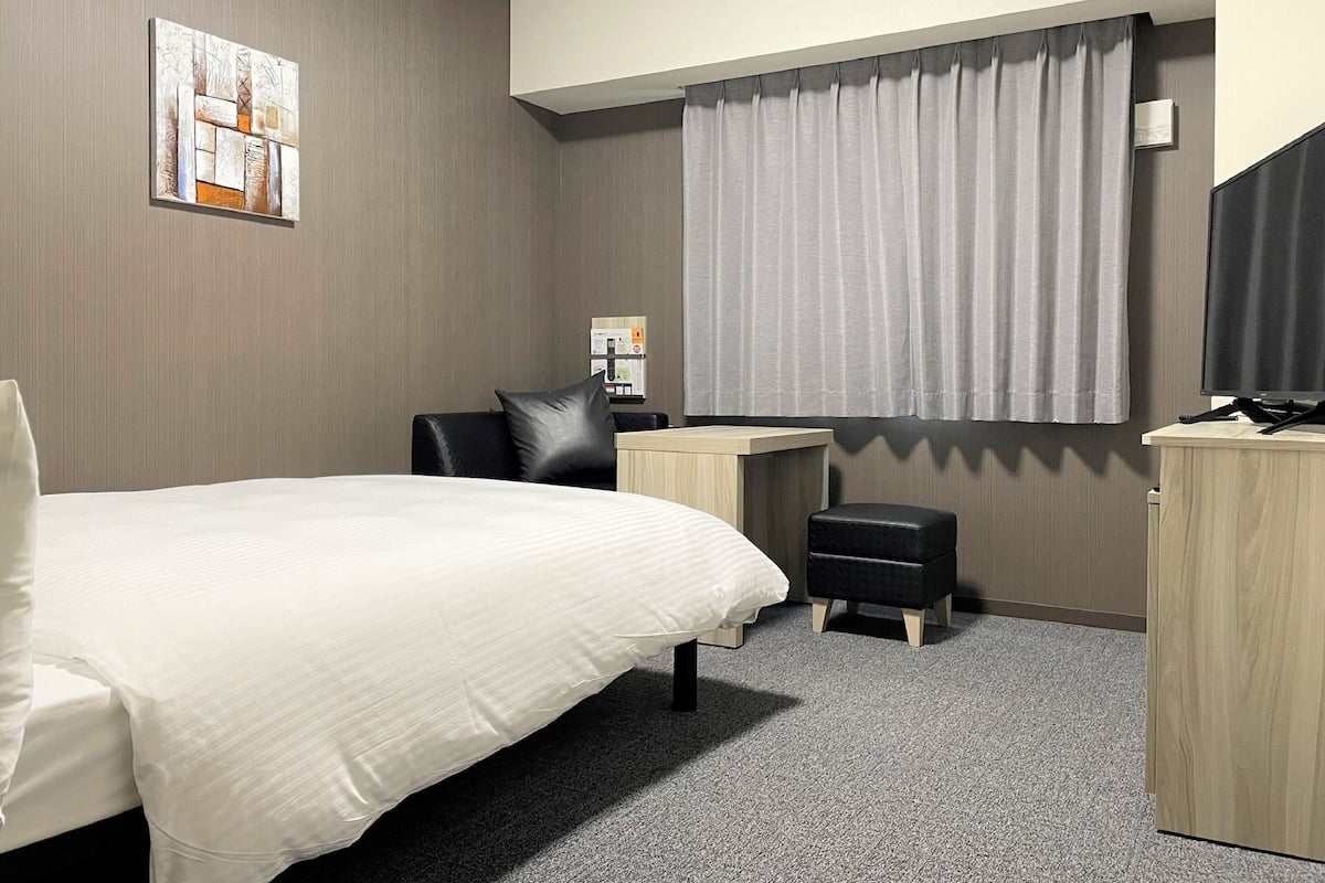A modern hotel room with a large bed, a desk, a chair, a small ottoman, and a television.