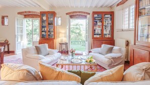 A cozy living room with two white sofas, a glass coffee table, and a brick fireplace. There are open doors leading to a garden, and a bookshelf filled with books is visible. - Villa Birdsong, among the Woods in Camaiore (Camaiore)