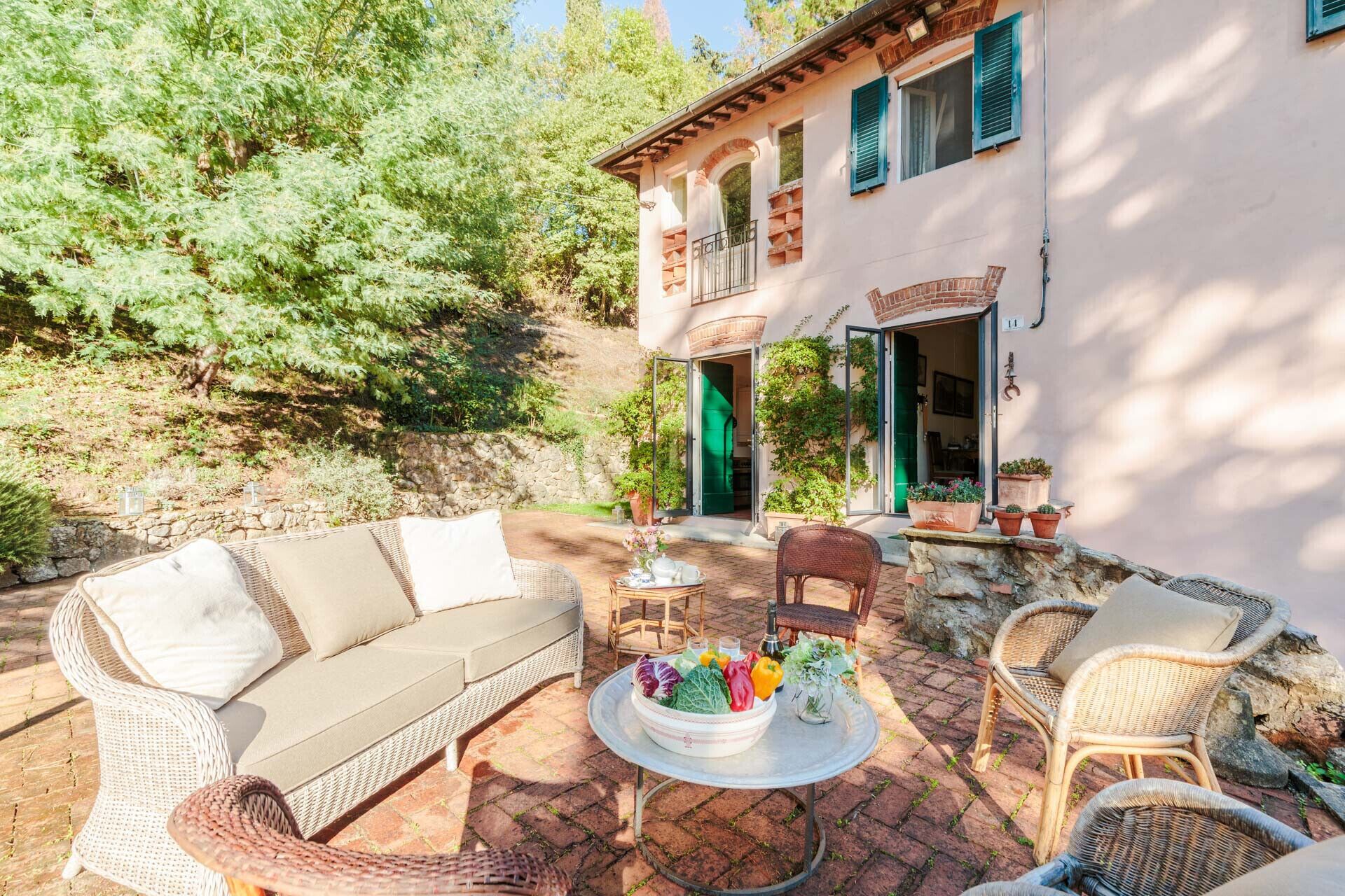 Un espacio al aire libre acogedor con mobiliario de mimbre, una mesa con un cuenco de verduras frescas y un edificio con pared de piedra y puerta verde.