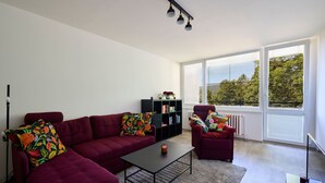 A modern living room with a red sofa, matching armchair, and a black coffee table. There is a bookshelf with books and decorative items, and a radiator under a window with blinds.