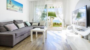 Een moderne woonkamer met een grijze bank, een salontafel en een televisie. Er is een groot raam met witte gordijnen, en er hangt een schilderij aan de muur.
