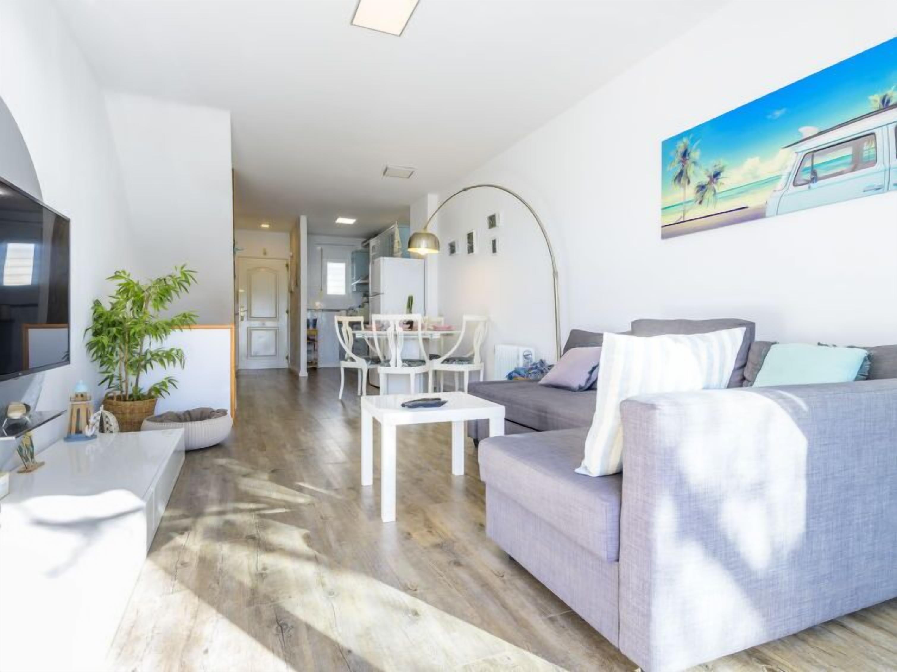 Un salon moderne avec un canapé gris, une table basse et un téléviseur. On y trouve également un coin repas avec une table et des chaises blanches. Une grande plante se trouve à gauche, et un tableau représentant un camion est accroché au mur.
