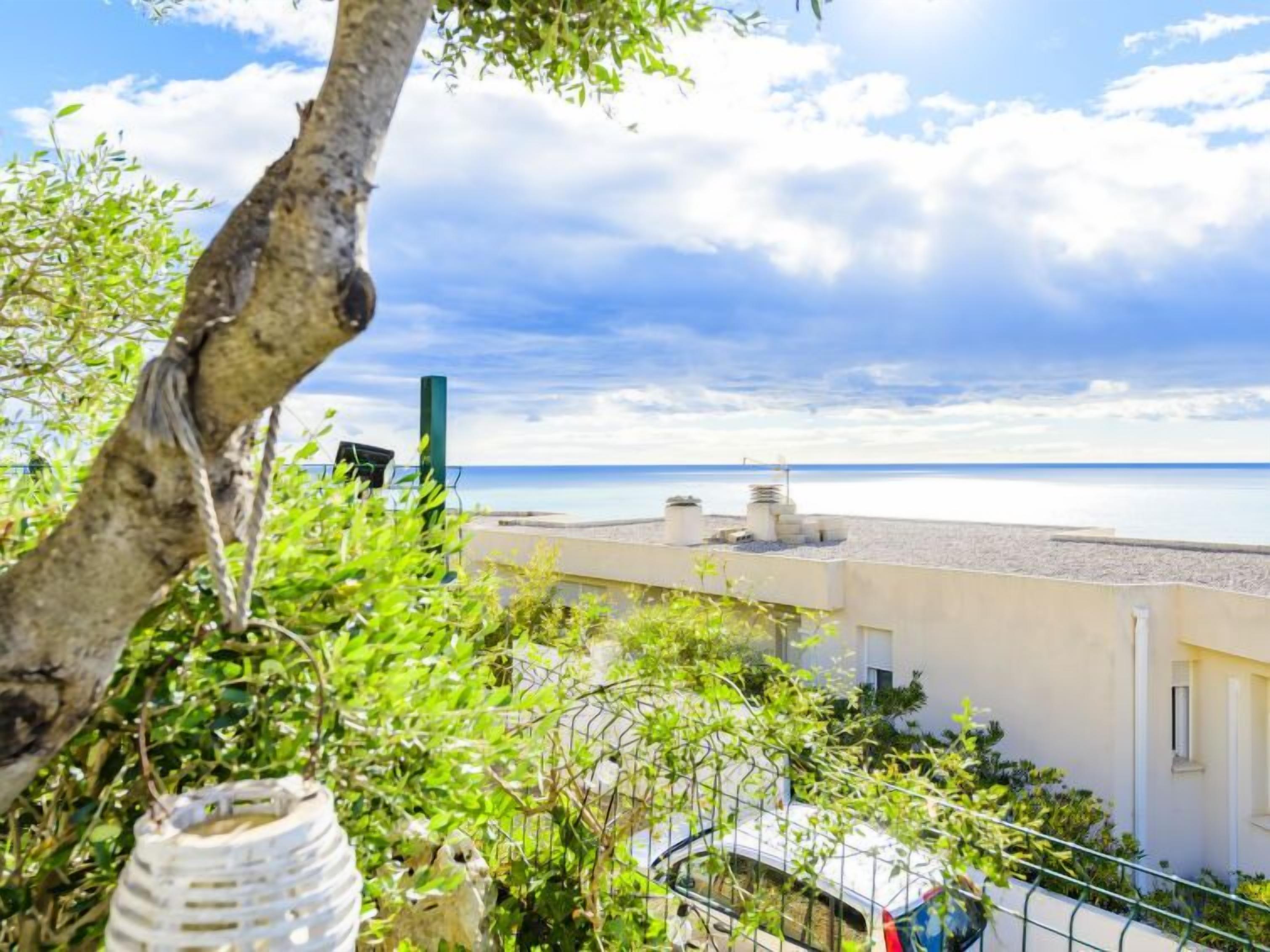 A balcony with a view of the sea, a tree, and a building with a white facade.