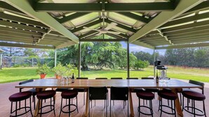 A covered outdoor dining area with a long table and multiple chairs, a ceiling fan, and a view of a grassy landscape. - Broke in the Vines - 6 bedroom farmhouse on working vineyard (Broke)