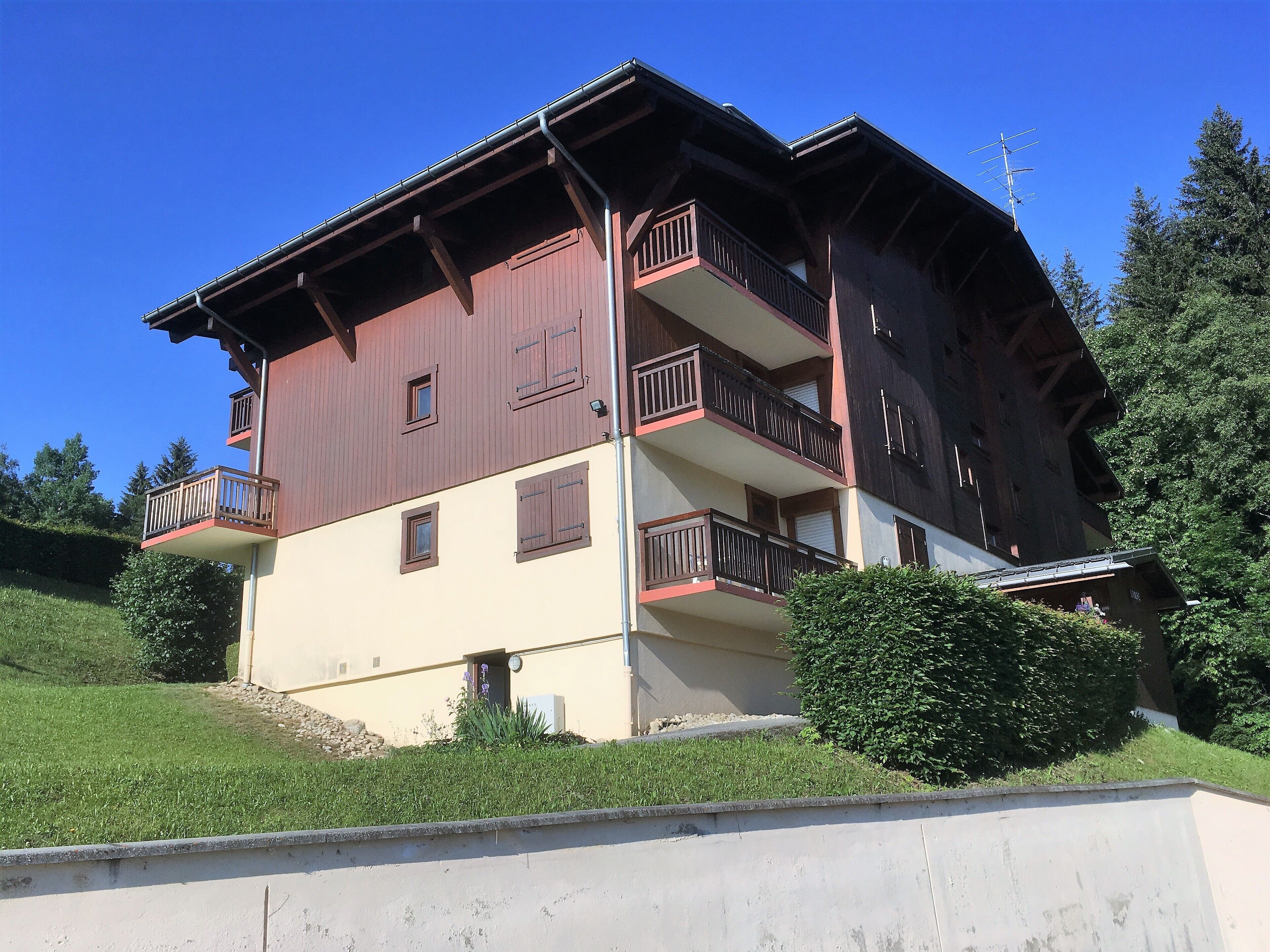 A multi-story building with balconies, a sloped roof, and a surrounding landscape.