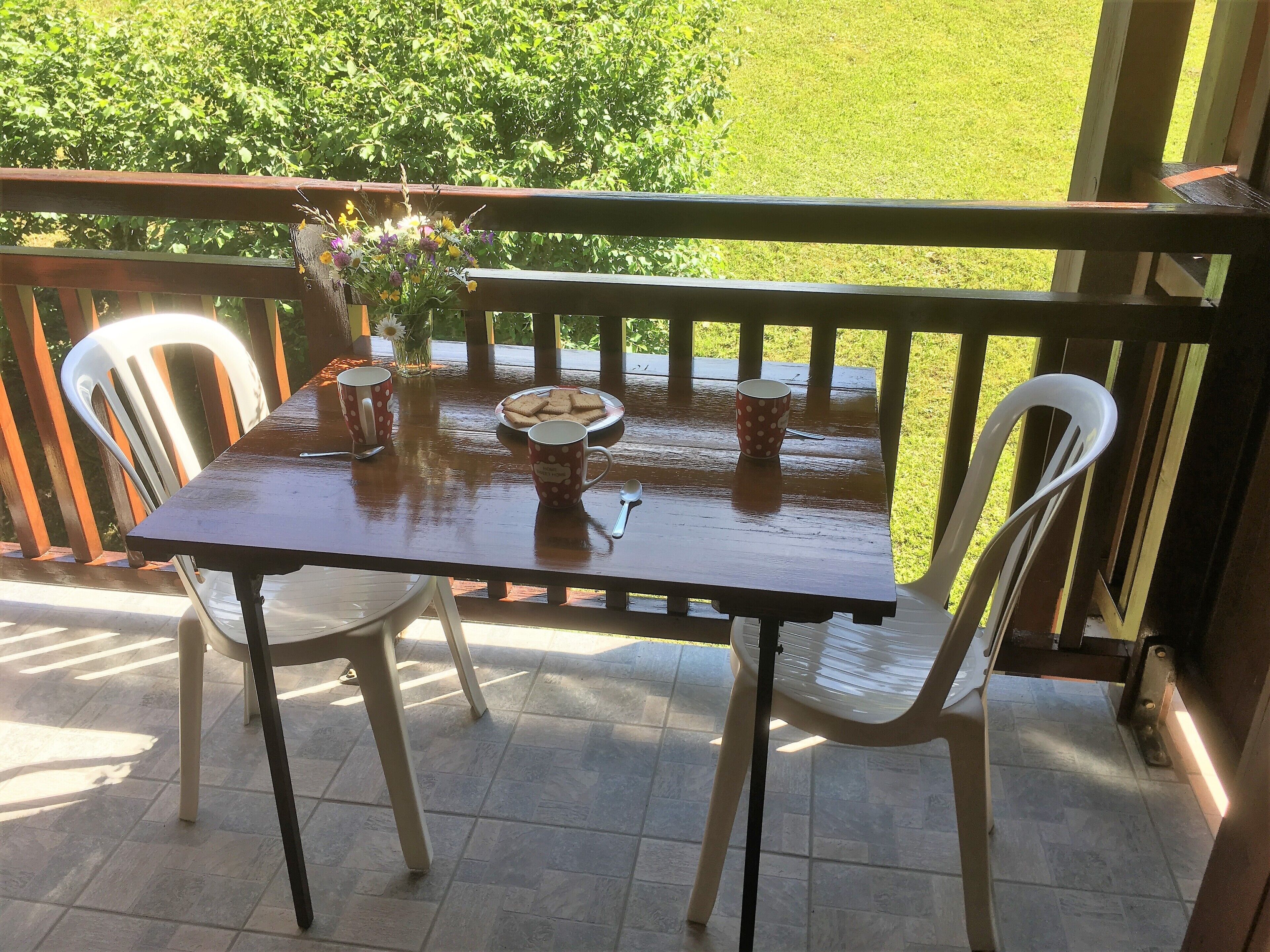 Ein Holztisch mit Stühlen auf einem Balkon, eine Vase mit Blumen und ein Teller Kekse.