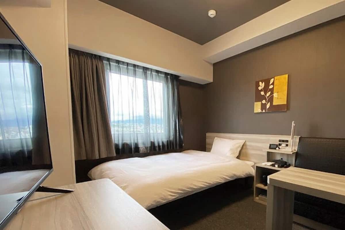 A hotel room with a bed, a desk, a chair, a bedside table, and a window with curtains.
