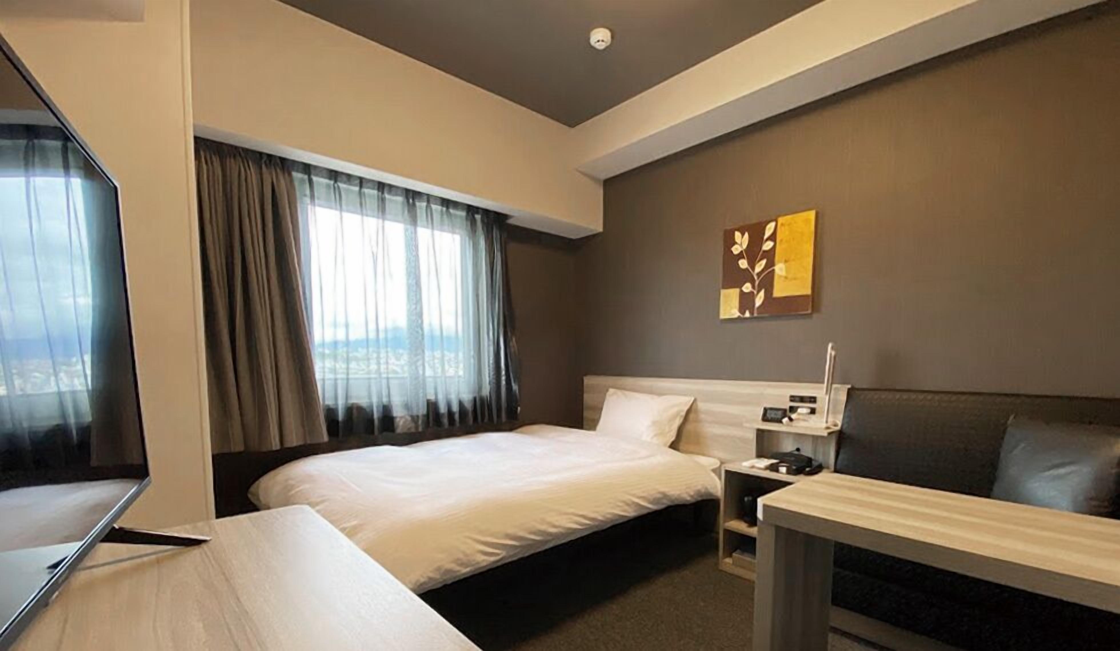 A hotel room with a bed, a desk, a chair, a nightstand, and a wall-mounted TV.