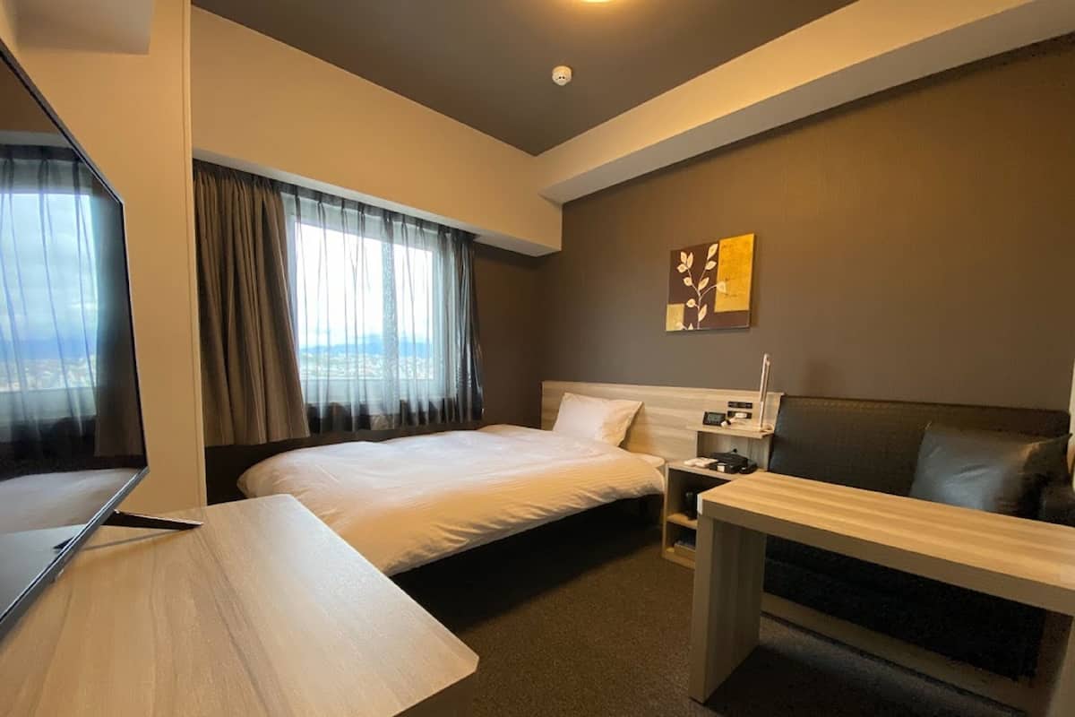 Habitación de hotel con cama, escritorio, silla, televisor y ventana con cortinas.