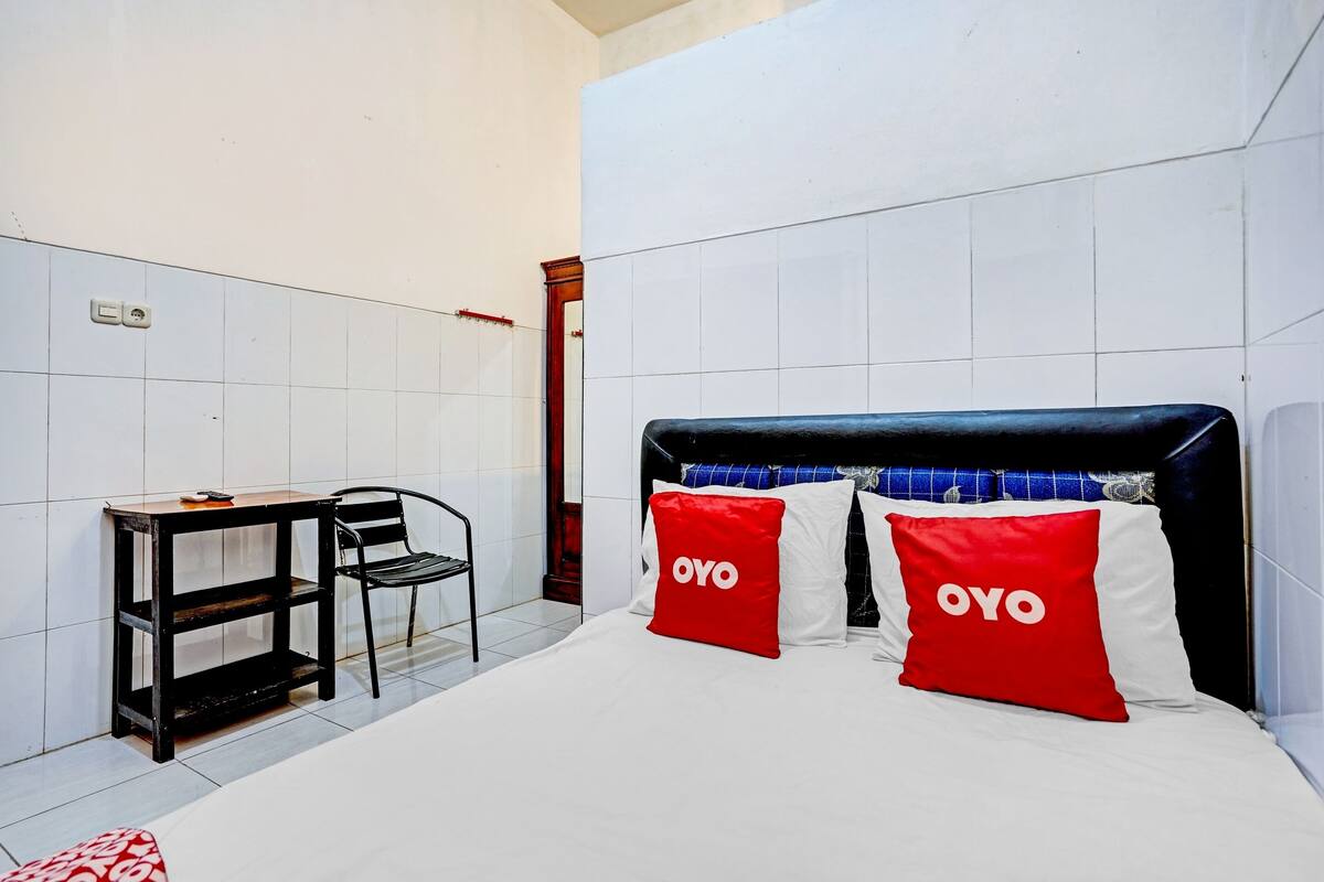 Hotelkamer met een bed, twee rode kussens met 'OYO'-branding, een houten nachtkastje, een stoel en een betegelde muur.
