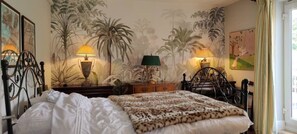 Une chambre à coucher avec un lit, deux tables de chevet, un chat et une fresque représentant des palmiers.